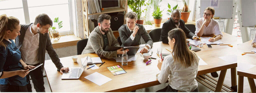 Employees at a conference room table having a discussion and reviewing materials on a laptop and papers.